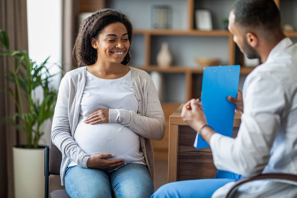 Vidám terhes fiatal fekete nő látogató orvos közel-keleti férfi a magánklinikán, terapeuta ad ajánlásokat boldog várandós hölgy. Egészségügyi ellátás a terhesség alatt koncepció - Fotó, kép