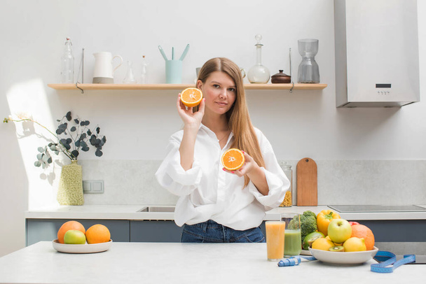 A young blond woman holds orange halves in her hands in the kitchen next to a table with fruits. The concept of diet, healthy eating. - Fotografie, Obrázek