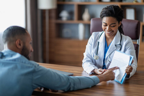 Smiling young black woman doctor showing middle eastern man patient treatment plan, arab guy attending therapist at newest private clinic, copy space, modern healthcare concept - Photo, Image