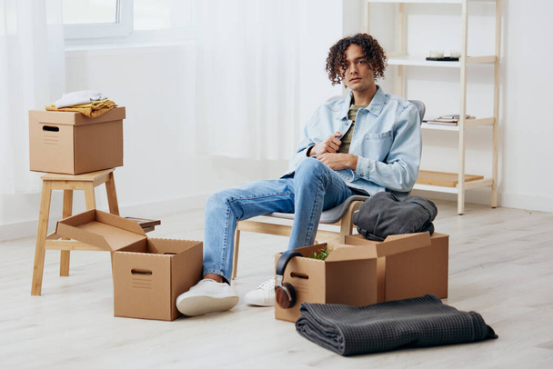 Όμορφος τύπος που κάθεται σε μια καρέκλα ξεπακετάρει με ένα κουτί στο χέρι και τακτοποιεί τα πράγματα. Υψηλής ποιότητας φωτογραφία - Φωτογραφία, εικόνα