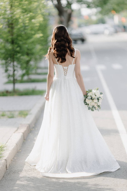 A stylish European bride in a white dress with a white bouquet is standing on the street. Photo from the back. High quality photo - Photo, Image