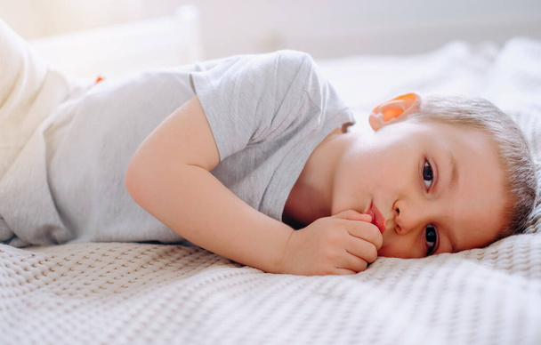 Крупный план маленького счастливого белого мальчика в серой футболке лежит на кровати и улыбается. - Фото, изображение