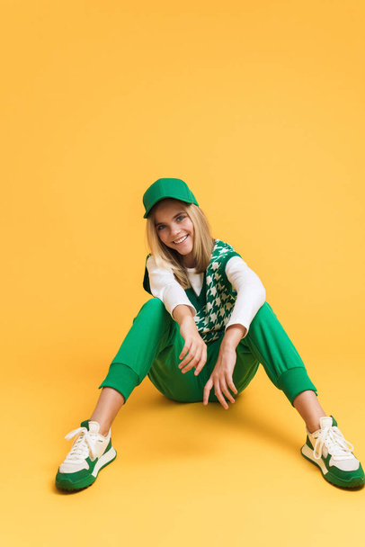 White girl wearing vest smiling and sitting on floor isolated over yellow background - Fotoğraf, Görsel