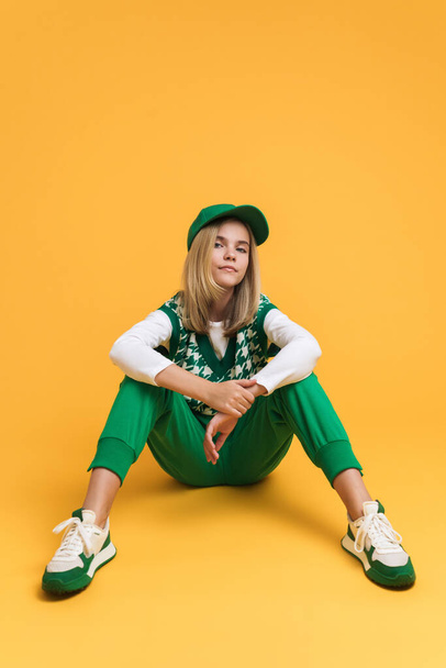 White girl wearing vest looking at camera and sitting on floor isolated over yellow background - Foto, afbeelding