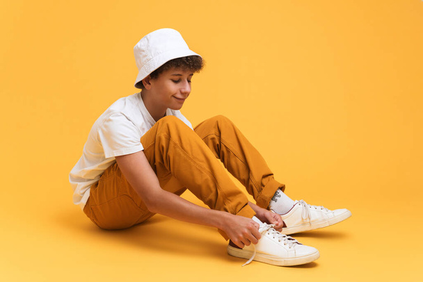White curly boy wearing panama smiling while sitting on floor isolated over yellow background - Photo, Image