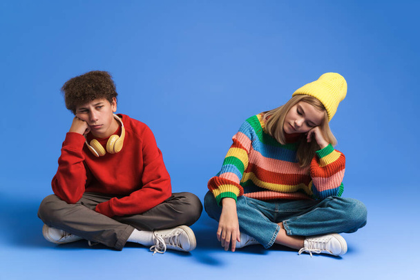 White sad girl and boy frowning while sitting on floor isolated over blue background - Foto, immagini