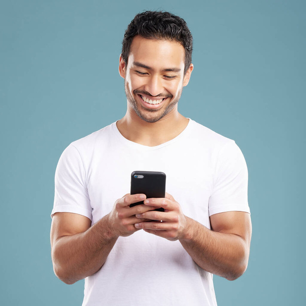 Handsome young mixed race man using his phone while standing in studio isolated against a blue background. Hispanic male sending a text message, using the internet online or browsing social media. - Foto, afbeelding