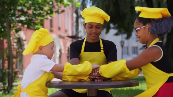 Командное построение многонациональных детей-поваров в шляпе шеф-повара и желтой форме фартука возлагают руки друг на друга, веселятся и смеются. Мультиэтническая активность детей в коммутации - Кадры, видео