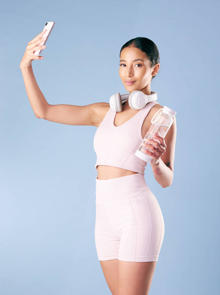 Mixed race fitness woman posing with her phone in studio against a blue background. Young hispanic female athlete taking selfie pictures with her smartphone to track her personal fitness growth. - Photo, Image