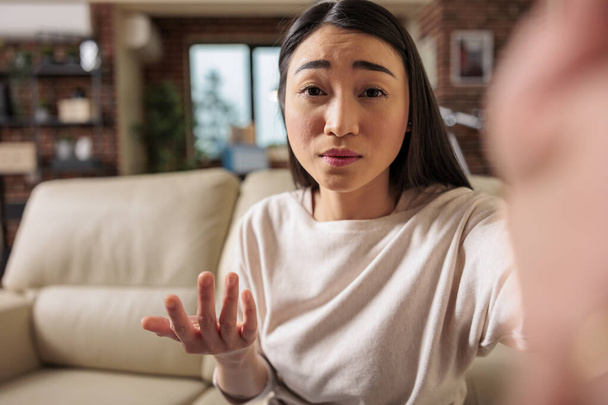 Красива автентична азіатська жінка-китайка за викликом у вітальні, тримає телефон, що відмовляється привітатися з камерою. Інтернет веб-технології дозвілля спілкування веб-камера чат відеодзвінки
 - Фото, зображення