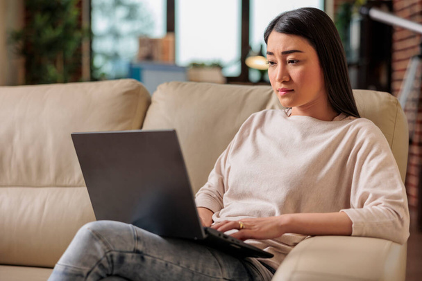Autentyczna azjatycka młoda kobieta korzystająca z laptopa, siedząca na kanapie w domu, piękna dziewczyna robiąca zakupy na czacie online w mediach społecznościowych, oglądająca film w wolnym czasie, wolny strzelec pracujący nad nowym projektem - Zdjęcie, obraz