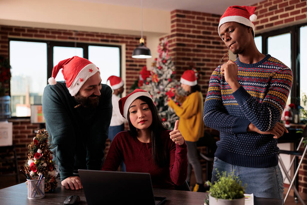 Multiethnic team of coworkers using laptop on christmas, working on project in office decorated with festive ornaments and xmas tree. People with santa hat doing teamwork during holiday. - Photo, Image