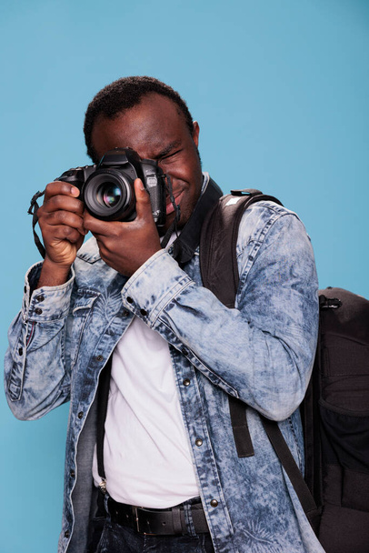 Confident photography enthusiast having photo device while taking picture. Young professional photographer with DSLR camera taking photo while standing on blue background. Studio shot - Fotoğraf, Görsel