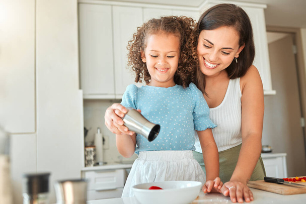 Happy mother teaching little daughter to cook in kitchen at home. Little girl adding seasoning to a bowl while making a salad with mom. - Фото, зображення
