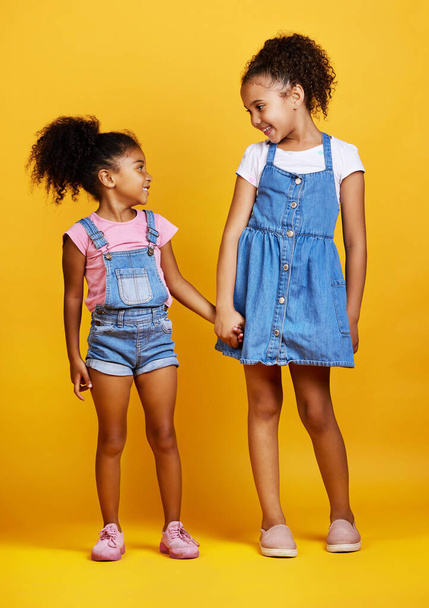 Mixed race girl sisters holding hands isolated against a yellow background. Cute hispanic children posing inside. Happy and carefree kids standing together. Sisters, siblings and best friends. - Foto, imagen