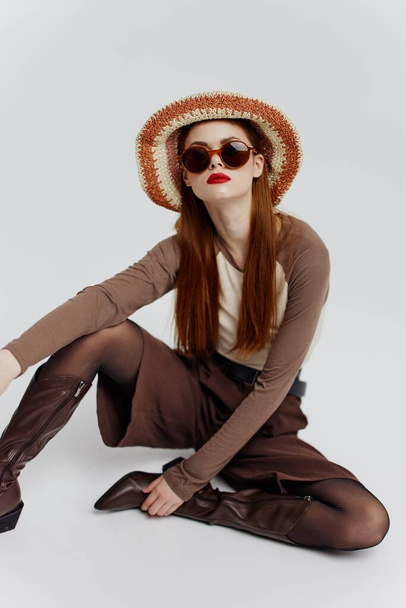 Een serieus jong vrouwelijk model met hoed, bril en laarzen zit op de grond in een witte studio. Modelcatalogus fotografie. Hoge kwaliteit foto - Foto, afbeelding