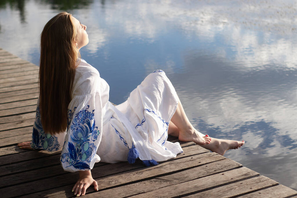 Девушка в вышитом национальном украинском костюме на пирсе на берегу озера. День независимости Украины, конституция, вышиванка. молодая женщина в синем платье на открытом воздухе. - Фото, изображение