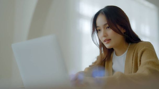 Посміхаючись азіатська молода жінка працює на ноутбуці в домашньому офісі. Молодий азіатський студент, використовуючи комп'ютерне дистанційне навчання, віртуальне навчання, електронне навчання, перегляд онлайн-освіти вебінар вдома
 - Фото, зображення