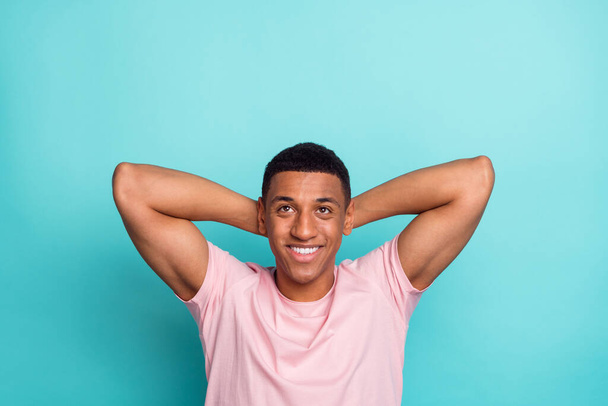 Photo of cheerful funny young man dressed pink t-shirt arms behind head looking up empty space isolated turquoise color background. - 写真・画像