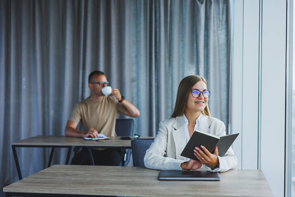 A business woman is holding a notepad and using a laptop while her European colleague is working in the background. Concept of modern successful women. Office workers sitting at desks in the office - Foto, imagen