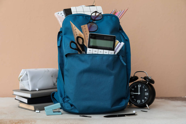 Шкільний рюкзак з канцелярськими приладами та будильник на столі на бежевому фоні
 - Фото, зображення