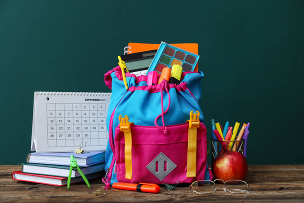 Школьный рюкзак с канцелярскими принадлежностями, календарь, яблоко и очки на столе на зеленом фоне - Фото, изображение