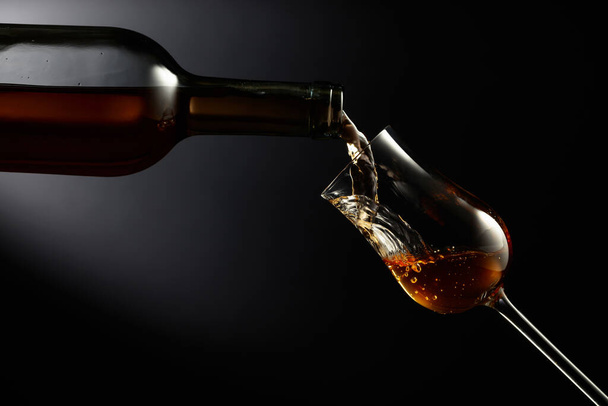 Glas premium alcohol op een zwarte achtergrond. Het drankje wordt uit een fles in een glas gegoten. Kopieerruimte. - Foto, afbeelding