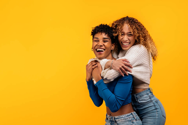 Stockfoto van vrolijke afro vrouwen knuffelen elkaar in studio schot tegen gele achtergrond. - Foto, afbeelding