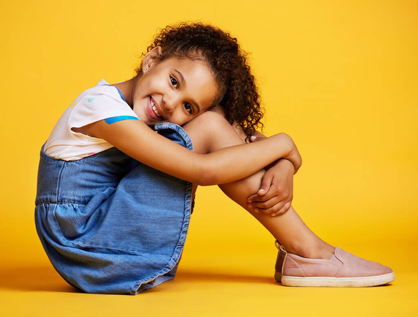 Studio portret mieszane rasy dziewczyna patrząc samotnie siedzi na żółtym tle. Słodkie latynoskie dziecko pozujące w środku. Szczęśliwy i ładny dzieciak uśmiechnięty i patrząc beztroski w ubraniach casual. - Zdjęcie, obraz