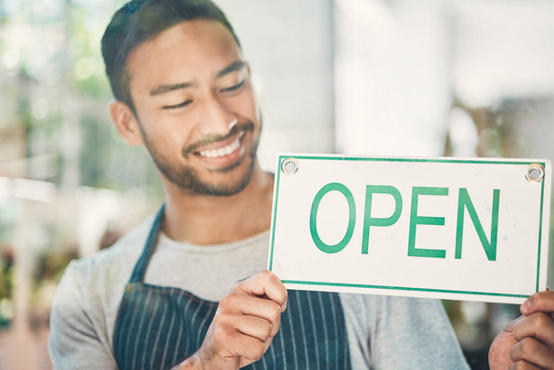 One young hispanic man hanging up an open sign in at a window on display in a cafe or store. Happy mixed race guy excited to open his shop and welcome customers. - Photo, image