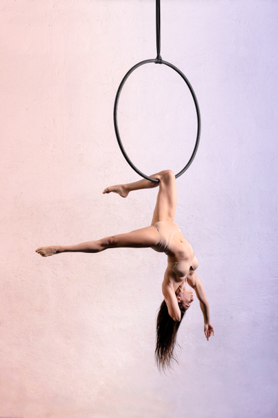 Full body side view of female gymnast hanging upside down from aerial hoop during training against light background in studio - 写真・画像
