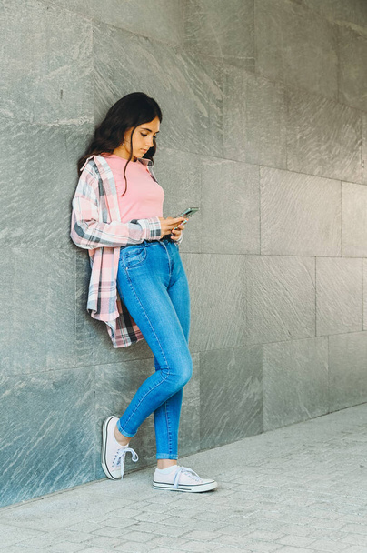Модна молода дівчина-підліток спілкується на своєму мобільному телефоні в соціальних мережах, коли вона стоїть, спираючись на сіру стіну з копійкою
 - Фото, зображення