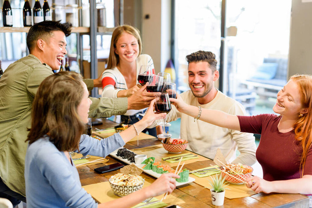 Group of cheerful multiracial friends looking at each other and clinking wineglasses with red wine while celebrating event together at table with food in restaurant - Foto, imagen