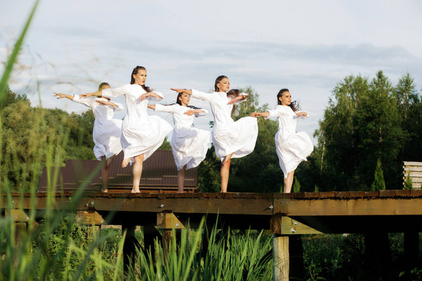 Ukrainian young girls in white dresses dance a stage dance in traditional style on a wooden pier against the background of nature and landscapes. - Foto, Imagen