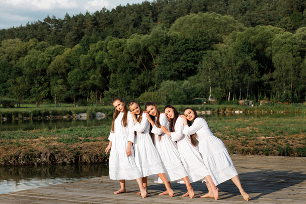 Украинские молодые девушки в белых платьях танцуют сценический танец в традиционном стиле на деревянном пирсе на фоне природы и пейзажей. - Фото, изображение