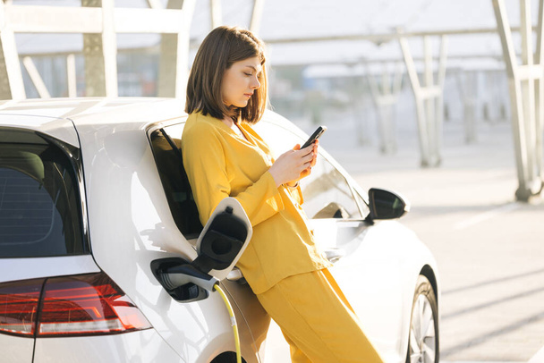 Caucasian woman stands with mobile phone near her electric car and waits when vehicle will charged. Charger plug of an electric car. Electric vehicle charging near an solar power plant - Foto, Imagem