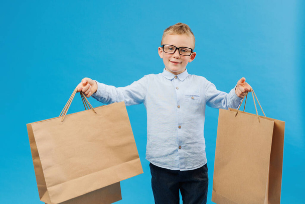 Cute funny boy in blue shirt holding paper bags isolated on blue background in studio. People lifestyle concept. The boy offers to go shopping - Photo, image