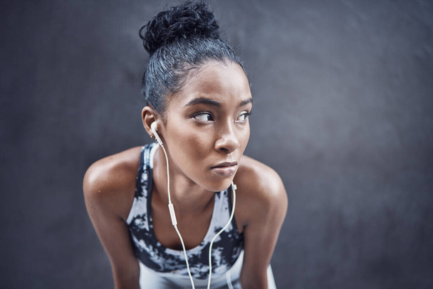 One active young mixed race woman wearing earphones and taking a rest break after run or jog exercise outdoors. Focused female athlete looking sweaty and tired but determined after challenging workout - Foto, Bild