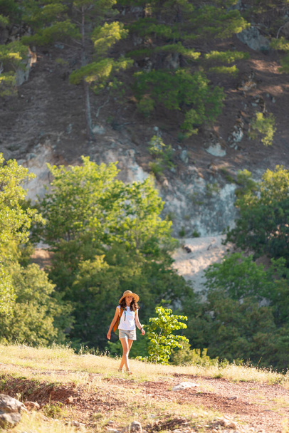 meisje reiziger met een rugzak gaat langs een bergpad. een jong meisje met een hoed met een rugzak wandelt langs de weg in het bos tegen de achtergrond van bergen. - Foto, afbeelding