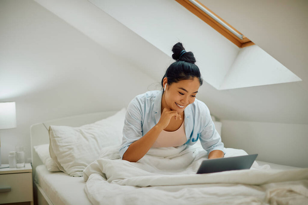 Mladá šťastná asijská žena pomocí počítače při relaxaci na posteli ve své ložnici. Kopírovat prostor.  - Fotografie, Obrázek