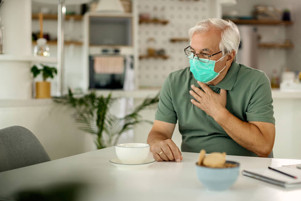 Senior man with a face mask coughing and holding his chest in pain at home.  - Foto, imagen