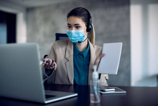 Young businesswoman explaining something during video call over laptop and wearing face mask due to coronavirus pandemic.  - Zdjęcie, obraz