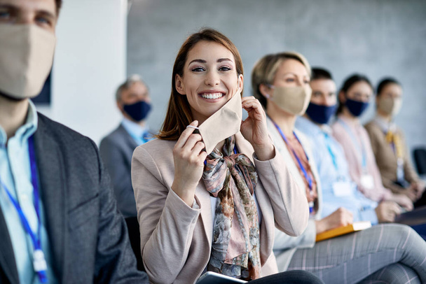 Νεαρή ευτυχισμένη επιχειρηματίας αφαιρώντας προστατευτική μάσκα προσώπου, ενώ παρακολουθεί ένα σεμινάριο στην αίθουσα συνεδρίων.  - Φωτογραφία, εικόνα