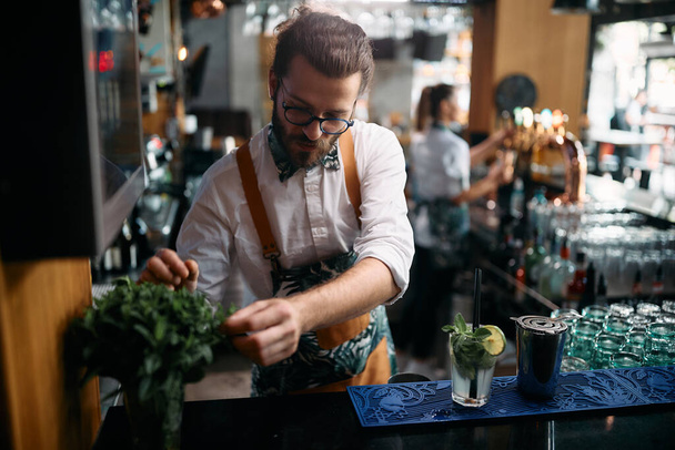 Young bartender using mint herb while preparing mojito cocktail at bar counter. - Fotó, kép