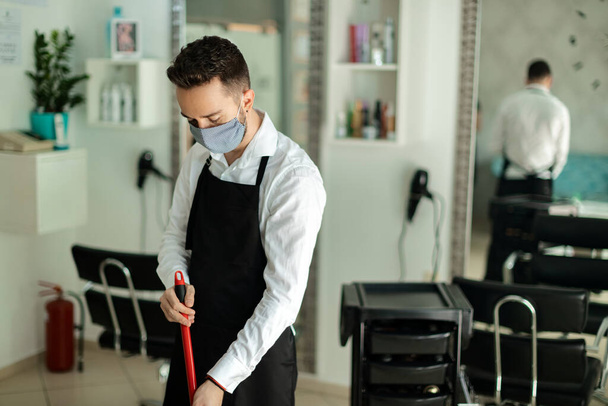 Молодой парикмахер в защитной маске и метле во время чистки салона после работы.  - Фото, изображение
