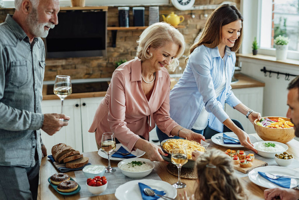 Szczęśliwej rozszerzonej rodziny korzystających w porze lunchu w jadalni. Kobiety podają jedzenie przy stole..  - Zdjęcie, obraz