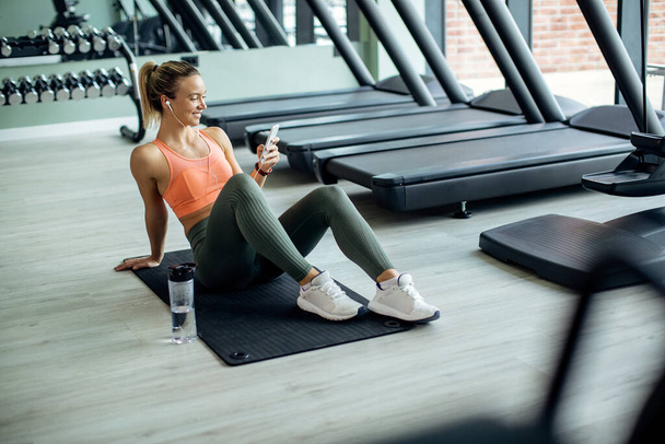 Happy female athlete taking a break from exercising and text messaging on mobile phone at health club.  - Valokuva, kuva
