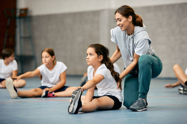 Маленькая девочка растягивается на полу и разогревается с помощью учителя физкультуры во время занятий в школьном спортзале.  - Фото, изображение