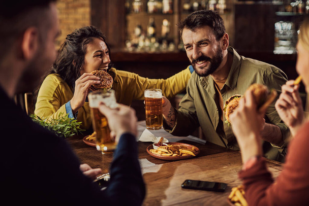 Счастливый человек веселится со своими друзьями, когда пьет пиво и ест бургеры в пабе.  - Фото, изображение