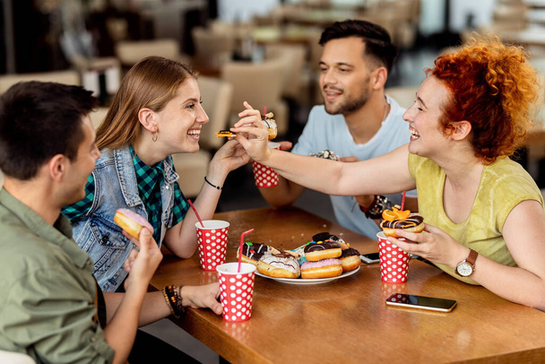 Group of carefree friends having fun while eating dessert in cafe. Focus is on women sharing a donut.  - Foto, imagen